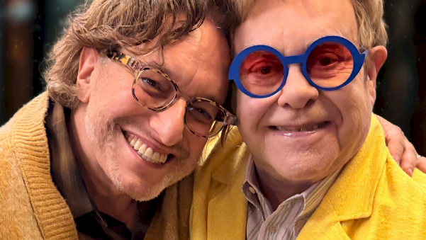 Two men smiling together, wearing bright yellow jackets; one with blue round glasses, posing closely.