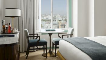 A hotel room with a bed, a small table and chairs by a window, a dresser with bottles, and a view of buildings outside, calm and modern.