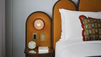 A hotel bed with a padded headboard, a small round clock and lamp on a wooden nightstand, and a white pillow with a decorative cushion.