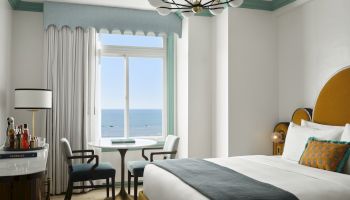 A bright hotel room with a round table by a large window overlooking the sea, a stylish bed, and modern chandelier, airy and inviting.