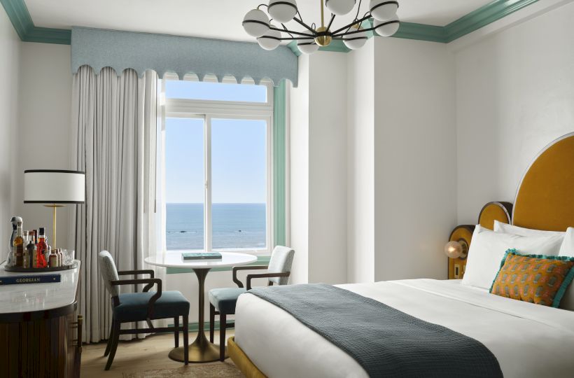 A bright hotel room with a round table by a large window overlooking the sea, a stylish bed, and modern chandelier, airy and inviting.