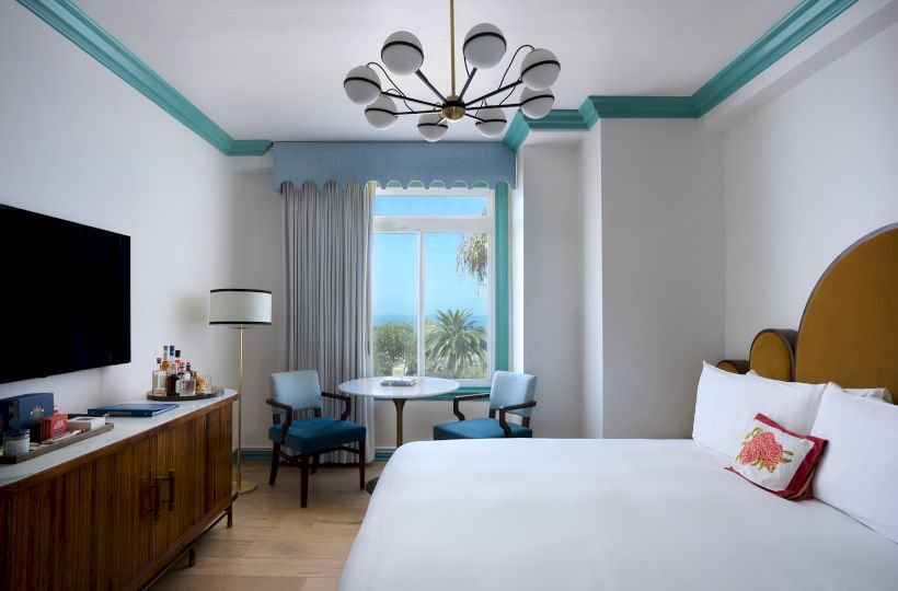 A bright hotel room with a teal-trim chandelier, small dining table, curtains, and a view through palm-framed windows.