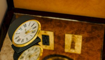 A small black clock with a white face sits on a mirrored surface, reflecting a wall with switches and a dim golden backdrop.