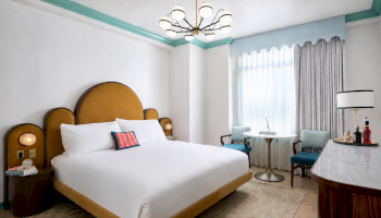 A modern hotel bedroom with a large bed, chandelier lights, teal accents and curtains, plus a small bar cart with bottles.