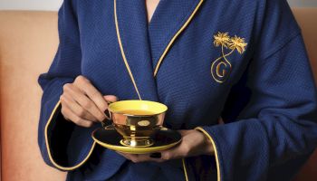 A person in a blue monogrammed robe holds a golden espresso/tea cup and saucer with hands over it.