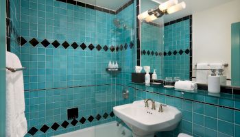 A bright bathroom with teal tiled shower, chrome rainhead, and a white sink and towels on a dark vanity.