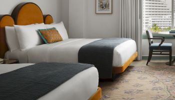 Two neatly made hotel beds with white linens, gray throws, and orange headboards in a bright room with a window and desk in the background.