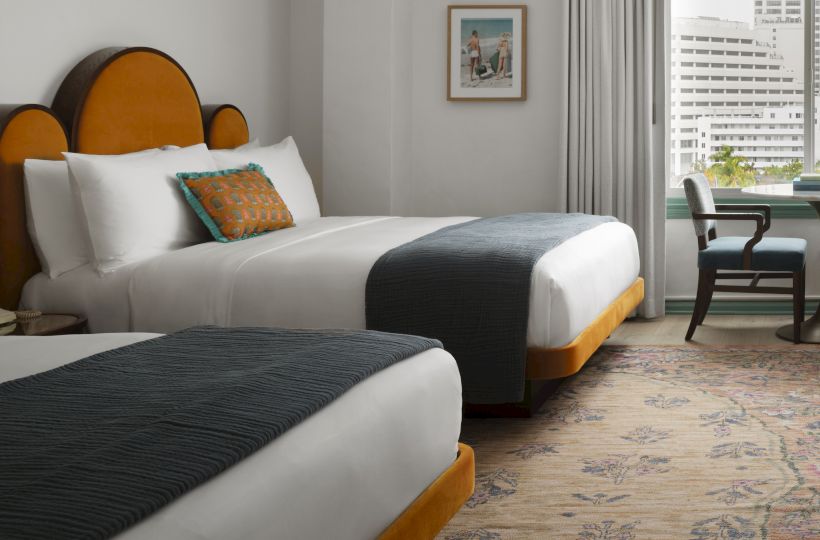 Two neatly made hotel beds with white linens, dark runners, orange-tinted headboards, a chair by a window, and a patterned rug in a bright room.