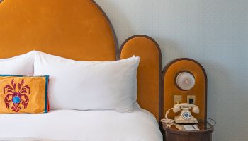 A hotel bed with white pillows and an orange throw sits beside a wooden nightstand with a vintage phone and lit lamp.
