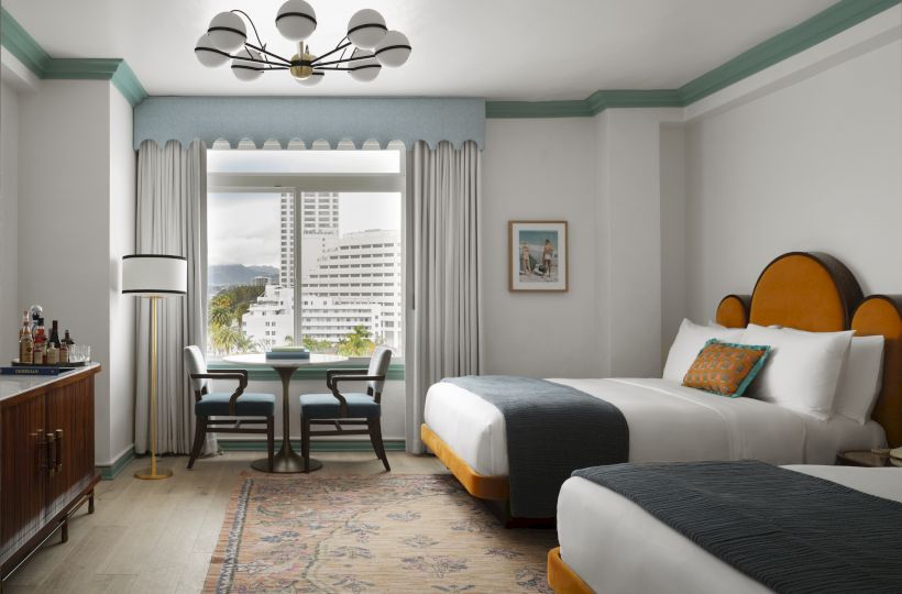 Bright hotel room with teal trim, chandelier, curtained windows, dining table, and a bed with colorful pillows.