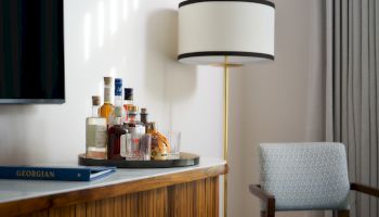 A minibar setup with several liquor bottles and glasses on a tray, beside a tall floor lamp in a modern room.