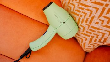 A mint-green hair dryer rests on an orange couch with a geometric-patterned orange-and-beige pillow beside it.