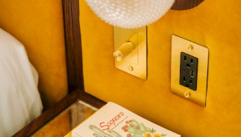 A small stack of books sits on a varnished nightstand beneath a textured globe lamp and warm yellow walls, next to a power outlet.