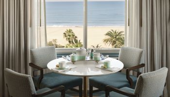 A round dining table set for four by a window view of a beach and palm trees; curtains frame the ocean scene beyond.