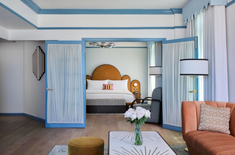 A bright hotel suite with an open blue door, plush bed, light curtains, pendant lights, and a sofa with cushions.