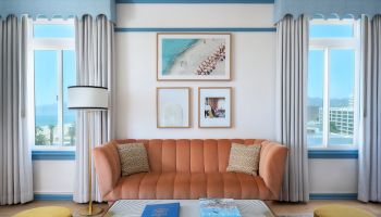 A bright living room with a tan sofa, gray curtains, large windows, and framed coastal artwork on the wall.