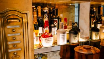 A cozy, vintage-style bar or apothecary shelf with bottles, wooden barrels, and brass fixtures, bathed in warm amber light.