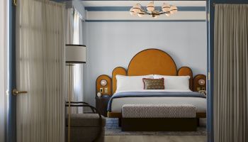 A hotel room with a large bed, blue trim, a curved headboard, two side tables, a bench at foot, and a sitting chair by a window.