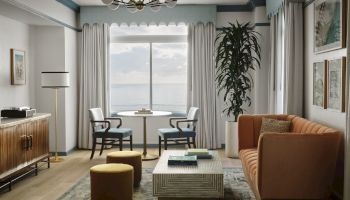 A stylish living room with a teal accent, large window, orange sofa, tall plant, striped curtains, and modern coffee table setup.