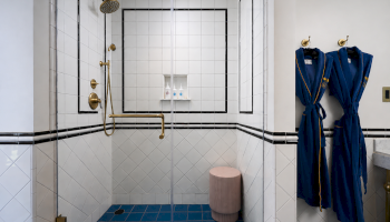 A modern tiled shower with gold fixtures and a rain head, plus blue bathrobes hanging on the wall and toiletries on a shelf.
