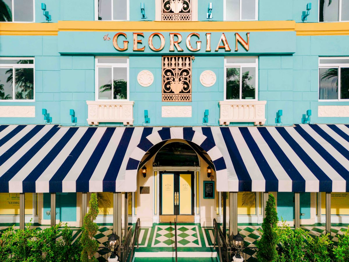 A pastel teal-blue building with a striped navy and white awning, arched doorway, tropical plants, and &ldquo;GEORGIAN&rdquo; sign above.