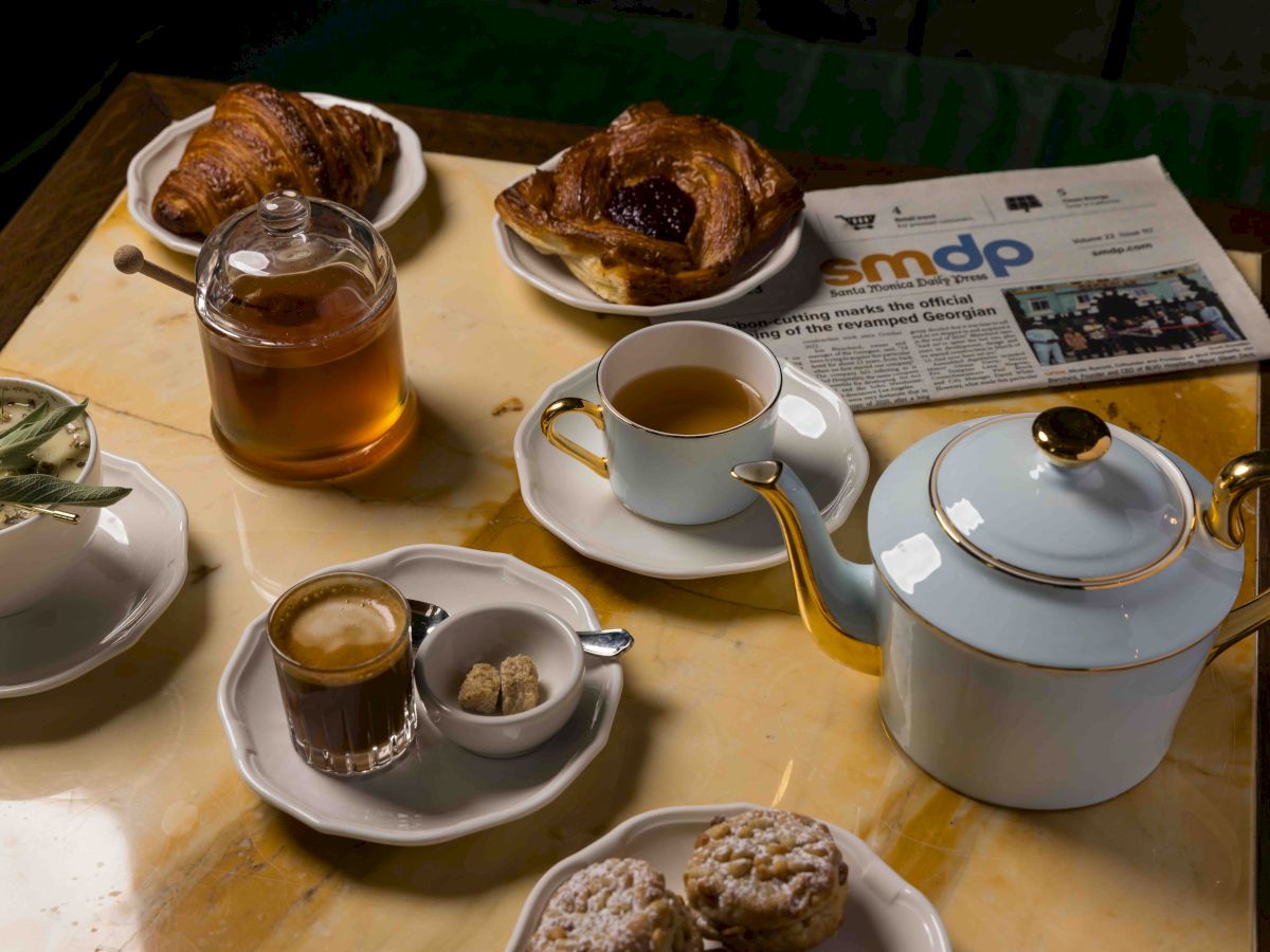 A table with tea, coffee, milk, pastries, croissants, a teapot, a cup of tea, a small sugar set, a newspaper, and muffins on a tray, all arranged neatly.
