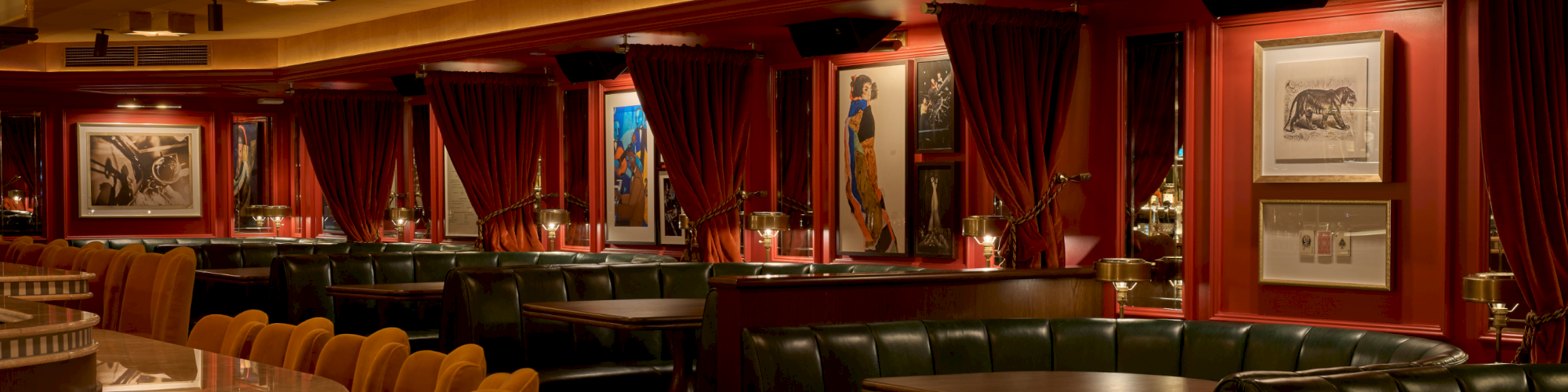 A dim, upscale lounge with red velvet curtains, leather sofas, and warm lighting; framed artlines and tables line the room like a cozy bar.
