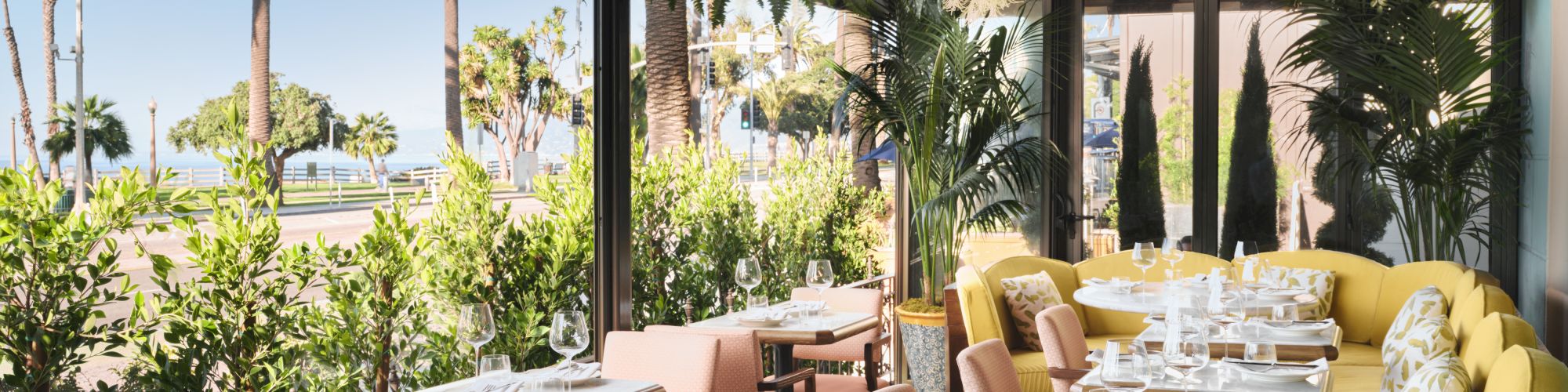 A stylish outdoor restaurant terrace with elegant tables, pink and yellow chairs, greenery, and hanging flowers under a shaded canopy.
