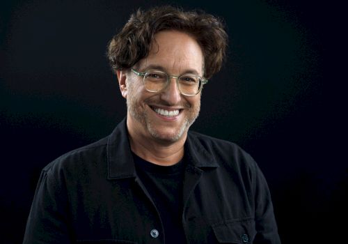 It shows a smiling man with glasses and dark hair, wearing a black jacket against a dark background, looks like a casual portrait.