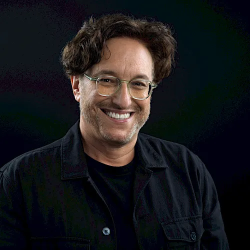It shows a smiling man with glasses and dark hair, wearing a black jacket against a dark background, looks like a casual portrait.