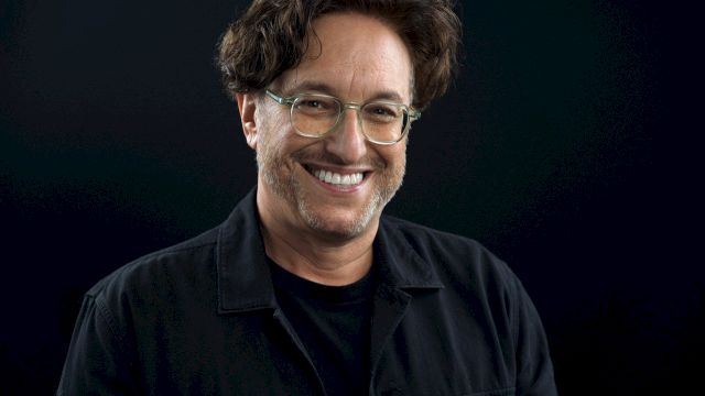 It shows a smiling man with glasses and dark hair, wearing a black jacket against a dark background, looks like a casual portrait.