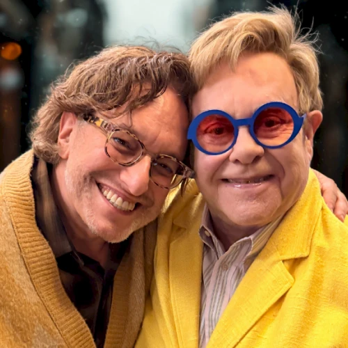 Two men smiling for a close-up photo; one in a mustard cardigan, the other in a bright yellow jacket with round blue sunglasses.