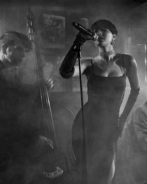 A moody black-and-white jazz trio: a singer at center with a microphone, a pianist on the right, and a guitarist/bassist on the left in smoky ambiance.