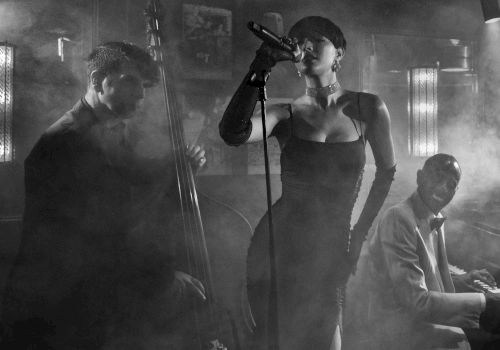 A moody black-and-white jazz trio: a singer at center with a microphone, a pianist on the right, and a guitarist/bassist on the left in smoky ambiance.