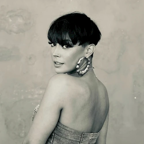 A stylish black-and-white portrait of a woman with a short haircut, off-shoulder dress, bold earrings, and a confident, sultry pose.