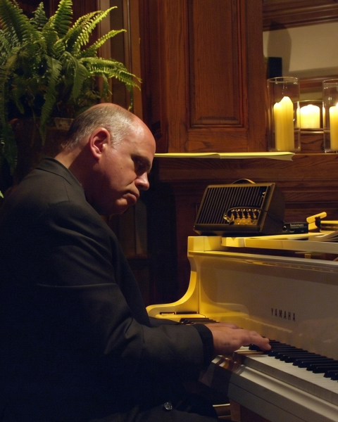 A man in a dark suit plays a grand piano in a warmly lit, wood-paneled room with plants and candles.