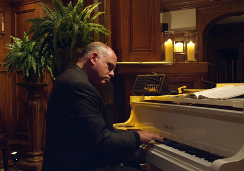 A man in a dark suit plays a grand piano in a warmly lit, wood-paneled room with plants and candles.