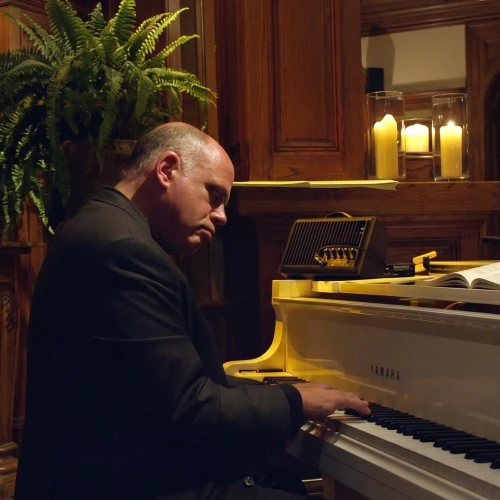 A man in a dark suit plays a grand piano in a warmly lit, wood-paneled room with plants and candles.