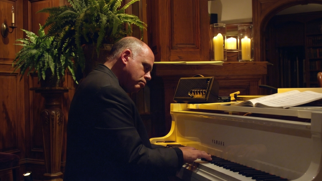 A man in a dark suit plays a grand piano in a warmly lit, wood-paneled room with plants and candles.