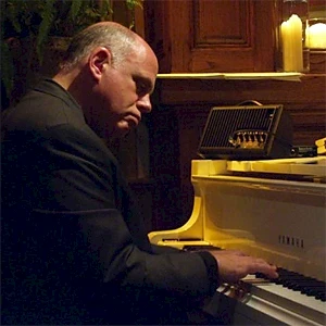 A man in a dark suit playing a white piano in a dimly lit room, focused on the keys.