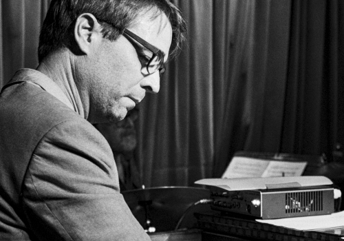 A man in a suit plays a piano/keyboard setup, focused, wearing glasses, vintage vibe, black-and-white scene.