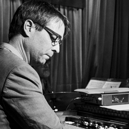 A man in a suit plays a piano/keyboard setup, focused, wearing glasses, vintage vibe, black-and-white scene.