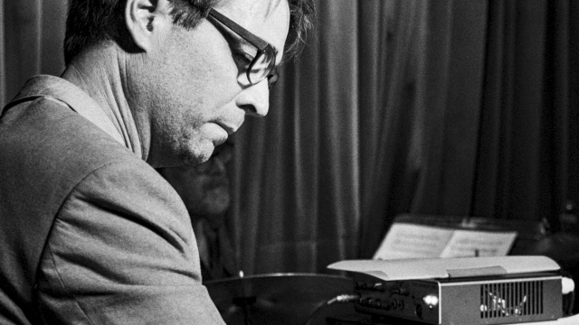 A man in a suit plays a piano/keyboard setup, focused, wearing glasses, vintage vibe, black-and-white scene.