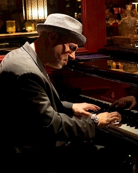 A man in a hat and sunglasses plays a piano in a dimly lit club, konzentred on the keys.