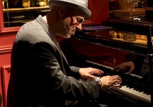 A man in a hat and sunglasses plays a piano in a dimly lit club, konzentred on the keys.