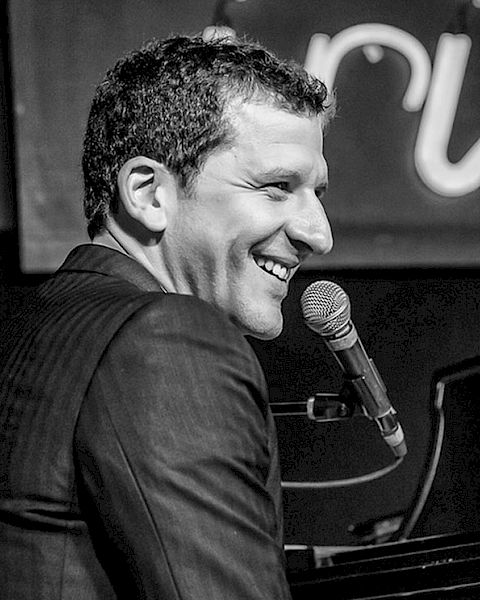 A smiling pianist singing into a mic at a piano, performing on stage.