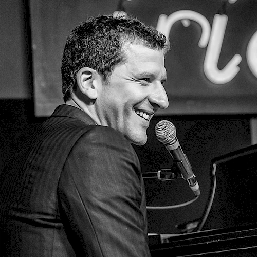 A smiling pianist singing into a mic at a piano, performing on stage.
