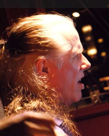 A man with long gray hair tied back sits in a dim room, eyes closed, lips parted as someone holds his shoulder in a supportive grip.
