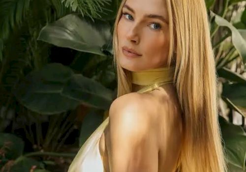 A woman with long blonde hair posing in a yellow top, standing among lush green tropical plants, looking over her shoulder.