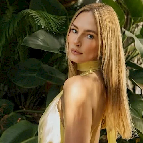 A woman with long blonde hair posing in a yellow top, standing among lush green tropical plants, looking over her shoulder.